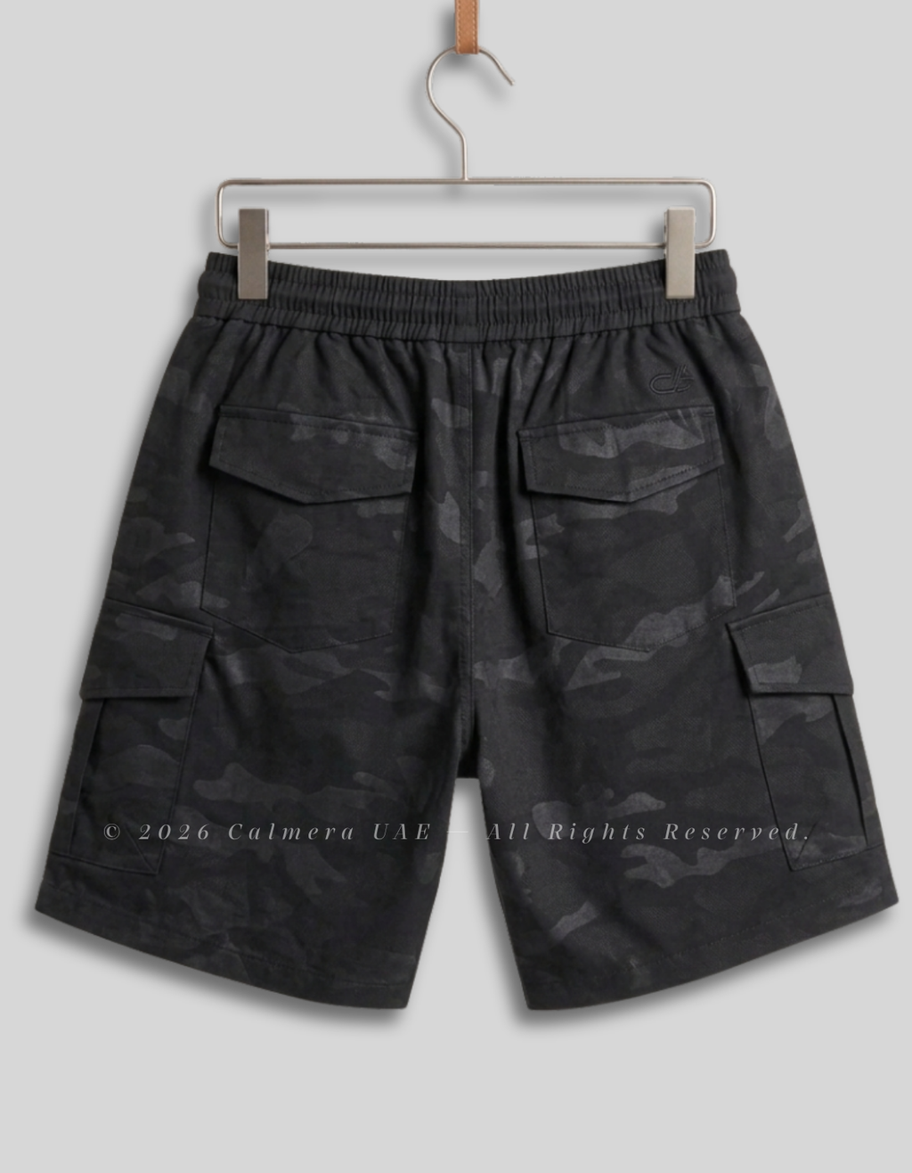 Black and dark gray camouflage cargo shorts with an elastic waistband, hanging on a metal hanger against a light gray background.