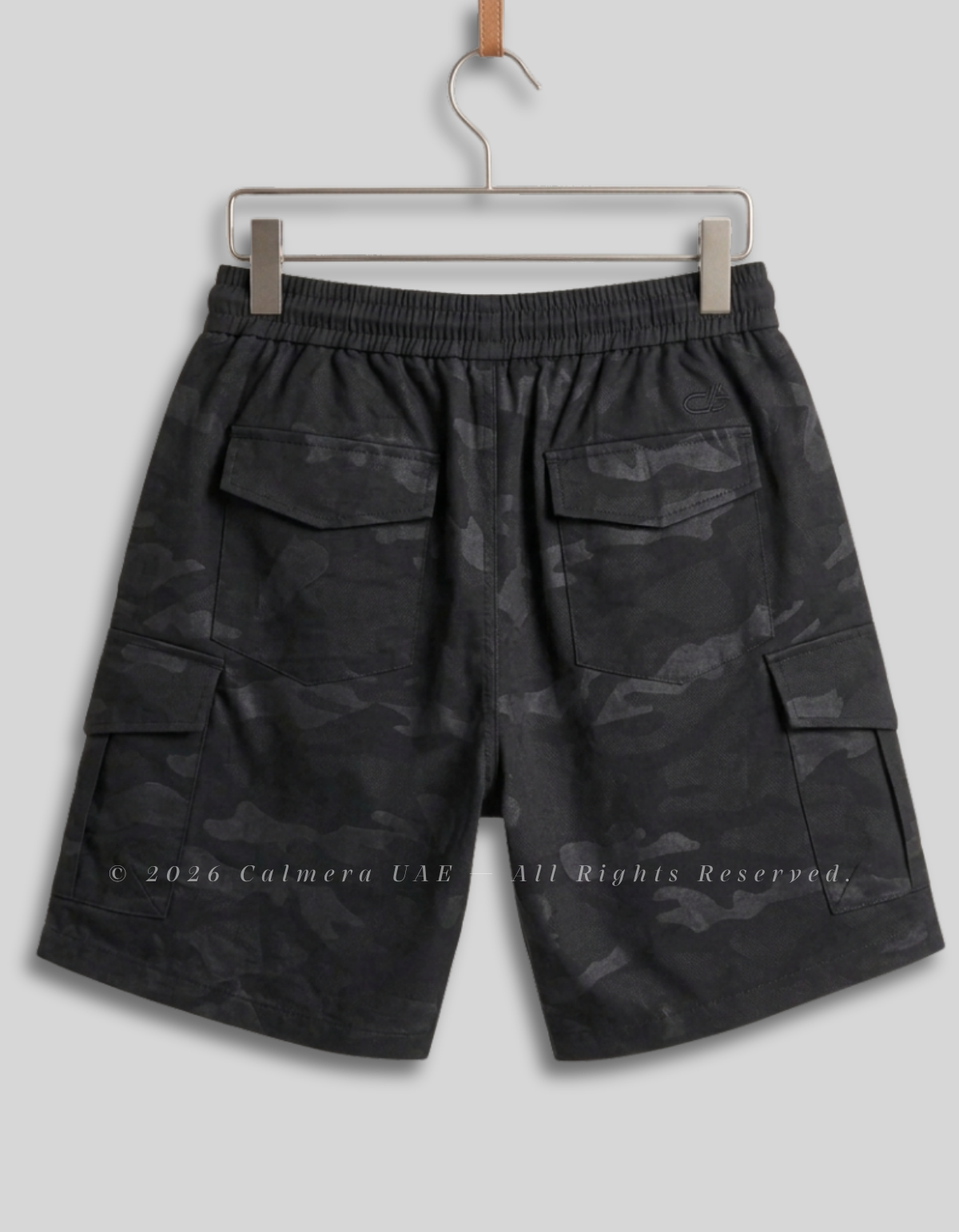 Black and dark gray camouflage cargo shorts with an elastic waistband, hanging on a metal hanger against a light gray background.