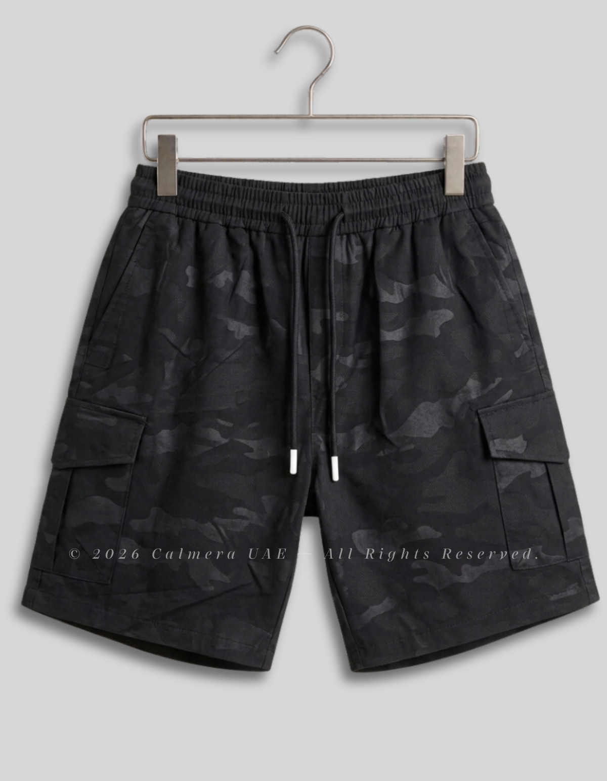 Black camouflage cargo shorts with an elastic drawstring waist, hanging on a metal hanger against a light gray background.