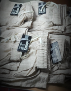 Multiple folded pairs of beige linen pants with elastic cuffs and drawstrings, each with a black and white 'Camelmet' size tag, stacked on a dark wooden surface.