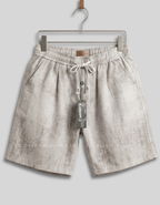 A pair of light beige linen drawstring shorts hanging on a metal hanger against a solid gray background. The shorts feature a textured, slightly mottled linen pattern, an elastic waistband, and a white drawstring.
