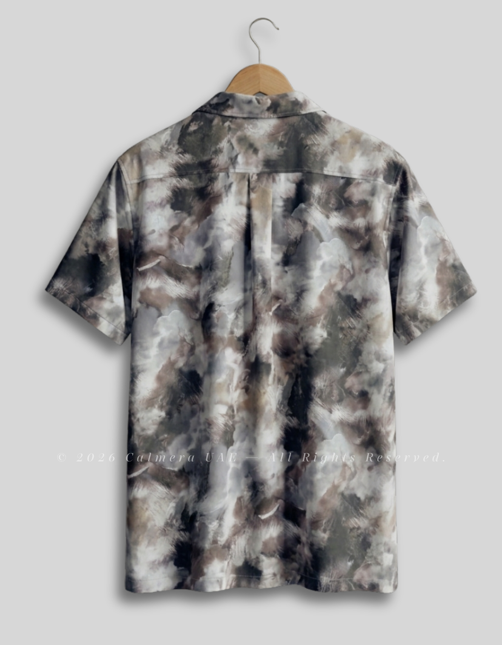 A short-sleeve button-up shirt with a brown, gray, and white tie-dye pattern, hanging on a wooden hanger against a light gray background.