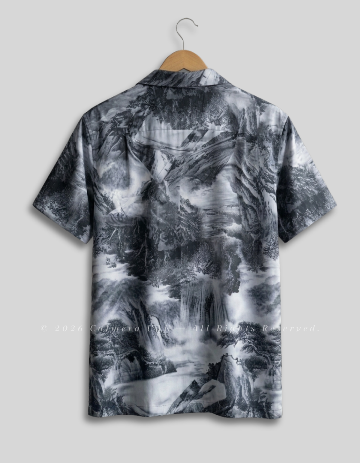 A short-sleeve camp collar button-up shirt with an all-over black, gray, and white misty mountain landscape print, hanging on a wooden hanger against a light gray background.