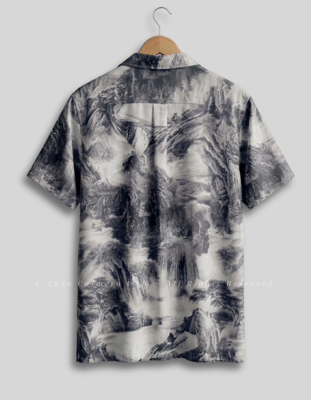 A short-sleeve button-up shirt hanging on a wooden hanger against a light gray background. The shirt features a monochrome, painterly landscape print with black, gray, and white tones depicting mountains, waterfalls, and mist.
