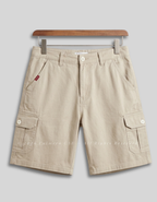 Beige cargo shorts with multiple flap pockets and button details, hanging from a wooden hanger against a light gray background.