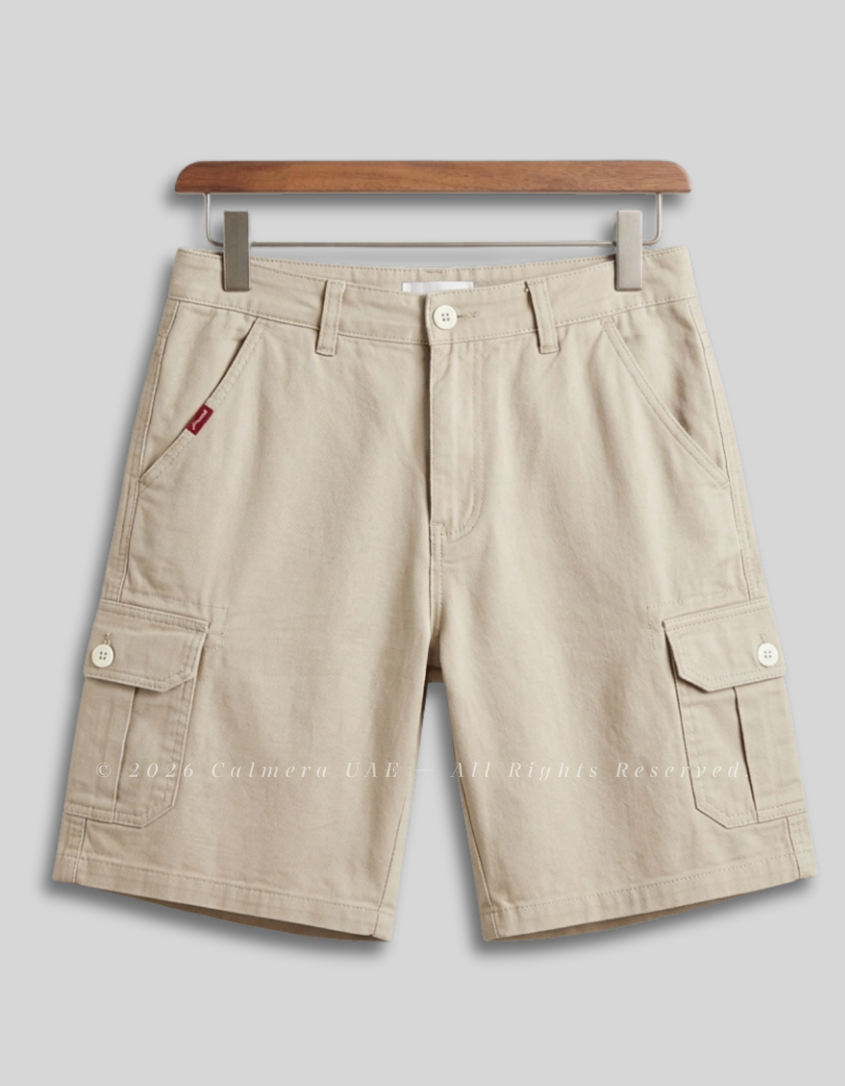 Beige cargo shorts with multiple flap pockets and button details, hanging from a wooden hanger against a light gray background.