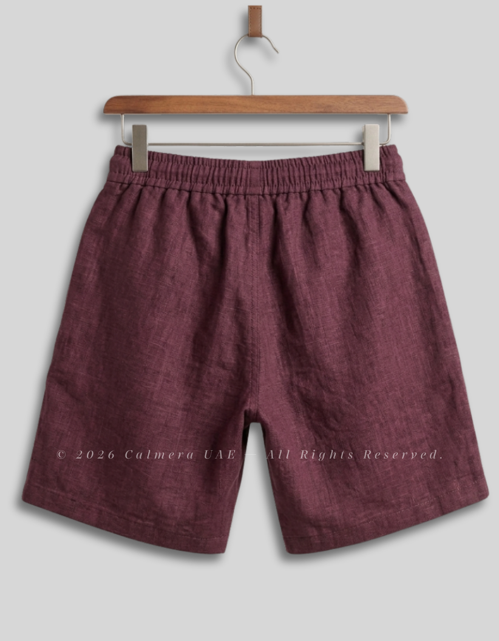 A pair of men's casual shorts in a textured burgundy fabric, hanging from a wooden hanger against a light gray background.