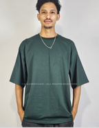 A man wearing an oversized dark green crewneck t-shirt, standing against a light gray background. The image features a watermark across the center reading 'CALMERA UAE - ALL RIGHTS RESERVED'.